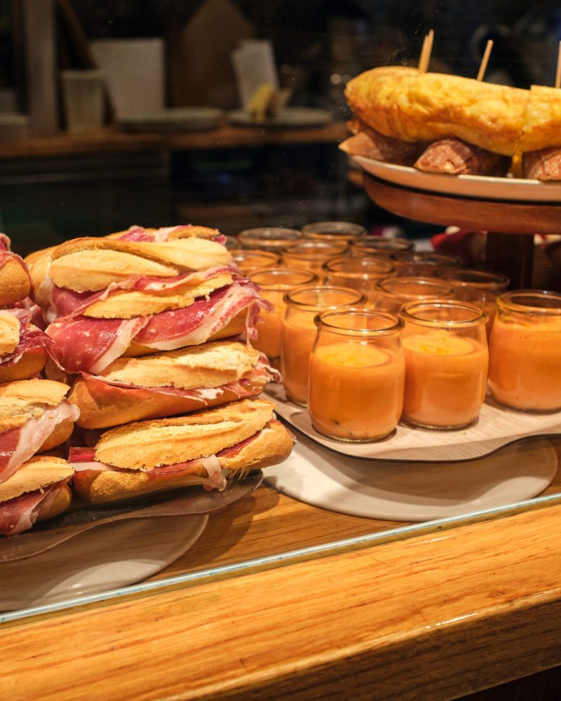 A variety of Spanish tapas, including ham and bread sandwiches and gazpacho soup, displayed on a countertop.